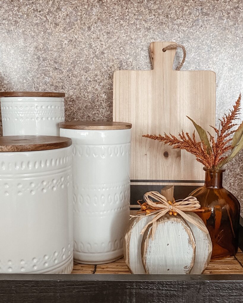 white canisters with wood tops sitting in a black and rattan serving tray with wood cutting board and fall decor